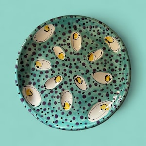 May include: A round, hand-painted ceramic plate with a turquoise background and a pattern of small purple dots. The plate features white, oval-shaped designs with yellow and black accents. The plate is a decorative item.