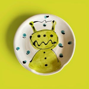 May include: A round white ceramic dish featuring a hand-painted lime green alien character with black outlines. The alien has three eyes, a jagged mouth, and two antennae. Small teal blue dots are scattered around the figure on the white surface.
