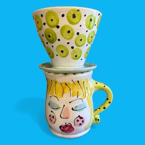 May include: A white ceramic mug with a black handle and the text "But First, Coffee" in a bold, black font. The mug is set against a blurred background, suggesting a cozy setting.