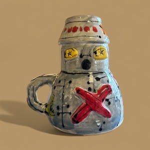 May include: A handmade ceramic mug in the shape of a robot. The mug is primarily gray with yellow star-shaped eyes, a red cross on the front, and a black dot for a nose. The handle is gray.