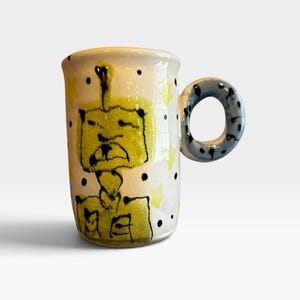 May include: A white ceramic mug featuring a hand-painted yellow figure with a square head and black outlines. The mug is decorated with small black dots and yellow star shapes across the surface. It includes a thick, round, blue-spotted handle.