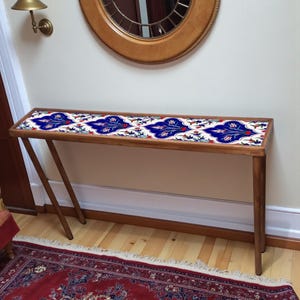 Op de afbeelding: Een houten console met een rechthoekig blad met een blauw en wit bloemen tegelontwerp. De tafel heeft schuine poten en staat tegen een neutrale muur met een ronde spiegel.