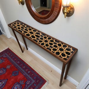 Könnte beinhalten: Ein langer, schmaler Holztisch mit einer Tischplatte aus schwarzen und goldenen geometrischen Fliesen. Der Tisch hat vier schlanke, abgewinkelte Beine und steht vor einer neutralen Wand. Ein roter Teppich ist auf dem Boden zu sehen.