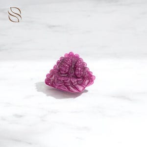 Könnte beinhalten: Eine geschnitzte, magentafarbene Edelstein-Skulptur mit zwei Figuren mit detaillierten Gesichtszügen und aufwendigen Kopfbedeckungen. Die Skulptur ist dreieckig und liegt auf einer weißen Oberfläche.