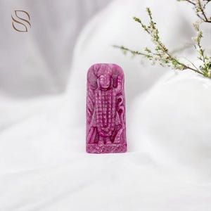 Puede incluir: Una escultura de piedra preciosa tallada de color magenta de una deidad. La figura se representa de pie con detalles intrincados, sobre un fondo blanco. La escultura es rectangular y presenta tallas detalladas de ropa y rasgos faciales.