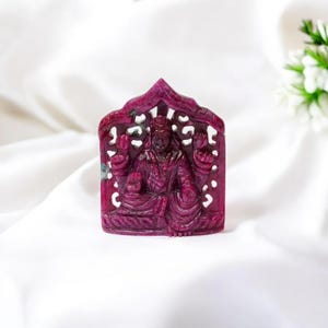 Puede incluir: Una escultura tallada en piedra preciosa de color rojo intenso que representa una deidad sentada. La figura está intrincadamente detallada, con múltiples brazos y ropa ornamentada. La escultura se encuentra sobre un fondo blanco, resaltando el rico color y detalle de la piedra preciosa.