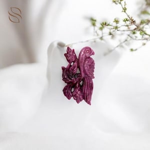 May include: A carved, magenta-colored bird pendant with intricate details, perched on a cluster of leaves. The pendant is set against a soft, white background, with a sprig of greenery in the upper right corner. The bird's feathers and the leaves are finely detailed.