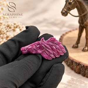 Puede incluir: Una piedra preciosa tallada con forma de cabeza de caballo, de color magenta intenso y rosa oscuro con un acabado pulido. La talla muestra texturas detalladas en la crin y en los rasgos faciales del animal.