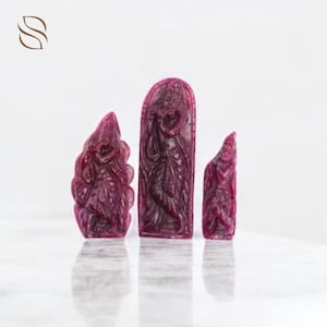 Könnte beinhalten: Drei geschnitzte Rubin-Skulpturen. Die Skulpturen sind tiefrot und zeigen Figuren in verschiedenen Posen. Die mittlere Skulptur ist ein hohes, rechteckiges Stück mit einer detaillierten Schnitzerei einer stehenden Figur. Die anderen beiden Skulpturen sind kleiner und haben abstraktere Formen.