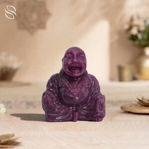Peut inclure: Une statue de Bouddha rieur, sculptée dans une pierre violet foncé. La statue a une surface lisse et polie et est représentée avec une large bouche ouverte. Le Bouddha porte une robe et est assis dans une pose détendue. L'arrière-plan est de couleur neutre.