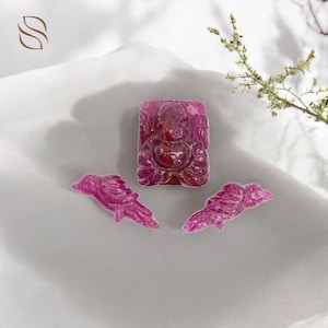May include: A collection of carved ruby gemstones. The set includes a rectangular Buddha pendant and two bird-shaped charms. The gemstones are a deep pink colour and are displayed on a white surface.