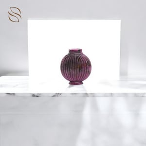 Könnte beinhalten: Eine kleine, runde, lila dekorative Vase mit vertikalen Rippendetails. Die Vase hat einen kurzen Hals und einen kleinen, kreisförmigen Sockel. Die Vase besteht aus einem steinartigen Material und steht auf einer weißen Oberfläche.