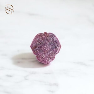 Puede incluir: Una gema tallada, de color púrpura oscuro, con un diseño floral ornamentado. La gema tiene forma de flor o hoja estilizada, con detalles intrincados y un acabado pulido. El fondo es una superficie de mármol blanco.