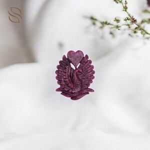 Puede incluir: Un colgante tallado en forma de corazón en tonos rojo oscuro. El colgante presenta un diseño de cisne con alas extendidas, un corazón en la parte superior y detalles intrincados de plumas. El fondo es una tela blanca suave.