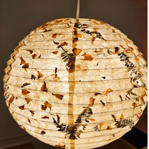 May include: A spherical paper lantern, illuminated from within. It features a design of dried leaves and botanical patterns in brown and orange hues. The lantern is suspended by a white cord.
