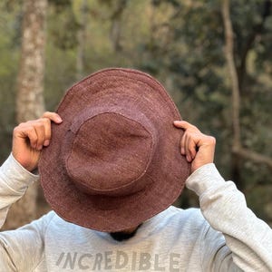 May include: A brown sun hat with a wide brim. The hat is made of a woven material. The person is wearing a grey sweatshirt with the word "INCREDIBLE" printed on it.