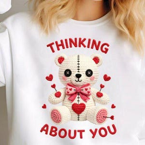 May include: White sweatshirt with a crochet teddy bear design. The bear has a pink bow, heart accents, and the text "THINKING ABOUT YOU" in red. The bear is cream-colored with black eyes.