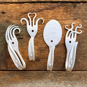 May include: Four silver-colored, repurposed silverware pieces arranged on a wooden surface. The collection includes two forks with unique curved tines, a spoon, and a fork with heart-shaped tines. Each piece has a floral design on the handle.