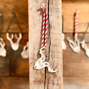 Puede incluir: Un adorno de reno plateado con una cuerda a rayas rojas y blancas cuelga de un poste de madera desgastada. El reno tiene un diseño estilizado con pequeños agujeros y un punto rojo. Otros adornos similares son visibles en el fondo.