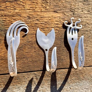 May include: Three handcrafted metal fork hooks mounted on a wooden surface. The hooks are made from repurposed silverware, with unique designs. The forks are bent and shaped into decorative hooks, showcasing a rustic, upcycled aesthetic.