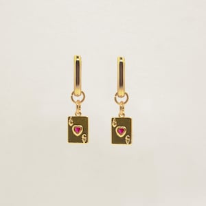 Op de afbeelding: Een paar goudkleurige oorringen. Elke oorring heeft een rechthoekige bedel met een rood hart en het nummer 6, dat op een speelkaart lijkt. De oorbellen zijn opgehangen aan een kleine gouden ring.