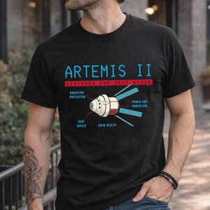 Puede incluir: Una camiseta negra con un gráfico de la nave espacial Artemis II. El texto dice ARTEMIS II, DESIGNED FOR DEEP SPACE, RADIATION PROTECTION, HEAT SHIELD, CREW HEALTH y POWER AND PROPULSON, con líneas que señalan partes específicas de la nave.