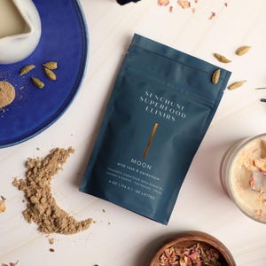 May include: A flat lay featuring a teal pouch of "Senchune Superfood Elixirs - Moon" with wild rose and cardamom. The image includes a blue plate with a small pitcher, a glass of drink with ice cubes and rose petals, and scattered ingredients.