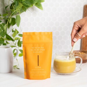May include: An orange pouch of SENCHUNE SUPERFOOD ELIXIRS, labeled "GLOW", sits next to a clear glass mug filled with a yellow beverage. A white pitcher and a wooden bowl are also present. A hand stirs the drink with a spoon.