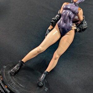 May include: A collectible figurine of a character with short lavender hair, wearing a glossy dark purple bodysuit, a black jacket, and black boots. The figure stands on a round black base with the text SECTION 9 TACTICAL printed on the front.