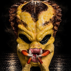 Könnte beinhalten: Eine detaillierte, gelbe Predator-Maske mit braunen Akzenten. Die Maske hat schwarze Augenhöhlen, scharfe weiße Zähne und rote und weiße Krallen. Die Maske ist eine Requisite aus dem Science-Fiction-Film.