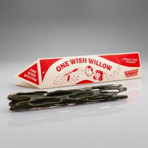 May include: A dark brown, porous, cylindrical wooden stick positioned in front of a triangular cardboard box. The box is cream and red with the text "ONE WISH WILLOW," "CRACK," "AMAZE YOUR FRIENDS!," and "You only get ONE WISH" printed on it.