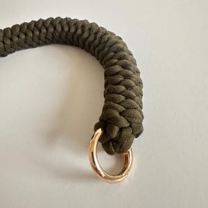 May include: Close-up of a braided olive green rope handle with a gold-colored metal ring. The rope is tightly woven, and the ring is attached to the end of the handle. The background is a plain white surface.