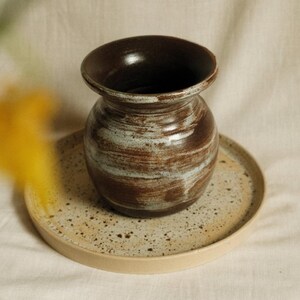 Op de afbeelding: Een handgemaakte keramische vaas met een donkerbruin interieur en een getextureerde, bruine en witte buitenkant. De vaas staat op een gespikkelde, ronde keramische schaal. De algehele esthetiek is rustiek en aards.