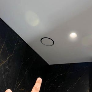 May include: A circular black recessed light fixture is installed flush into a smooth white ceiling. The ceiling meets a wall covered in dark black marble tiles featuring thin, light-coloured veining patterns throughout the stone surface.