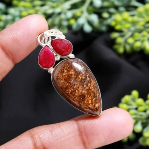 May include: A silver pendant featuring a large, teardrop-shaped brown stone with gold flecks, accented by two red ruby gemstones. The pendant is held by a finger against a blurred background.