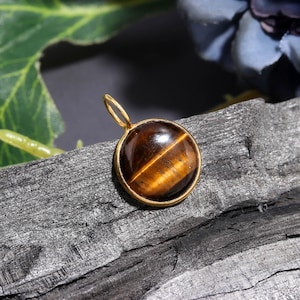 Puede incluir: Colgante redondo con una piedra ojo de tigre, engastada en un marco y una argolla dorados. La piedra es marrón con bandas amarillo dorado. El colgante se presenta sobre una superficie oscura y texturizada, para joyería o piedras preciosas.