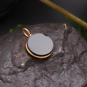 May include: A gold pendant featuring a circular, black gemstone. The pendant has a gold loop for a chain. The stone is smooth and polished, reflecting light. The pendant is set against a dark, textured background, giving a contrast to the gold.