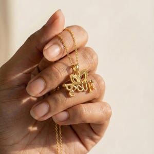 Può includere: Una collana con pendente dorato con un design di scrittura dettagliato. Il pendente è sospeso a una catena sottile. La collana è tenuta in mano su uno sfondo morbido e neutro. Il design del pendente è intricato.