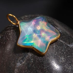Puede incluir: Un colgante en forma de estrella con un marco y una fianza dorados. El colgante tiene una superficie brillante e iridiscente con una gama de colores, incluyendo azul, verde y rosa. El colgante está sobre un fondo oscuro y texturizado.