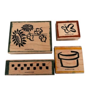 以下が含まれることがあります： 黒インクのデザインが施された4つの木製スタンプ。1つのスタンプはヒマワリ、もう1つは蝶、3つ目は植木鉢、最後は一連の小さな長方形です。各スタンプは木製のベースと緑色のボーダーが付いています。