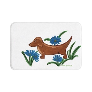 May include: A rectangular white bath mat featuring a brown dachshund illustration centered among blue flowers and green leaves. The text created by Brandi is printed in small green letters in the lower right corner of the mat surface.