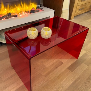 Könnte beinhalten: Ein rechteckiger Couchtisch aus durchscheinendem rotem Acryl mit Wasserfallkanten-Design. Zwei kugelförmige, goldfarbene Mosaikglas-Kerzenhalter mit warmer Innenbeleuchtung stehen auf der glatten, reflektierenden Oberfläche des Tisches.