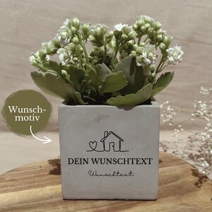 Könnte beinhalten: Ein quadratischer grauer Betonübertopf mit einer kleinen weiß blühenden Pflanze. Auf der Vorderseite befindet sich eine schwarze Strichzeichnung eines Hauses mit Herz sowie der Text "DEIN WUNSCHTEXT" und "Wunschtext". Ein grüner Kreis mit "Wunsch-motiv" ist zu sehen.