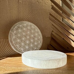Puede incluir: Dos placas circulares de cristal de selenita blanca con un diseño geométrico de flor de la vida grabado en la superficie. Una placa está en posición vertical, la otra está plana. Las placas están sobre una superficie de madera.