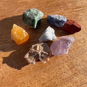 Könnte beinhalten: Eine Auswahl an rohen Edelsteinen in verschiedenen Farben, darunter Grün, Orange, Blau, Rot und Lila. Die Steine liegen auf einer Holzoberfläche und zeigen ihre natürlichen Texturen und Farbtöne. Eine Sammlung von Kristallen.