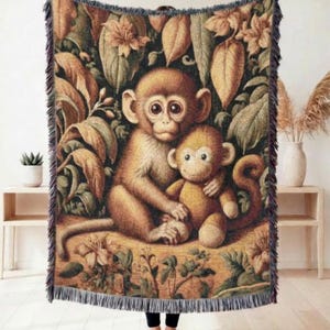 May include: A woven throw blanket featuring a whimsical illustration of two monkeys amidst lush foliage. The blanket showcases a warm color palette of browns, greens, and tans, with a fringed edge. The design evokes a cozy, nature-inspired theme.