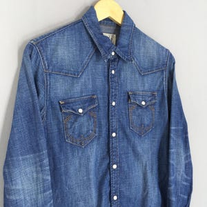 May include: A blue denim shirt with long sleeves, two chest pockets, and a button-down front. The shirt has a classic western-style yoke design and is hanging on a wooden hanger.