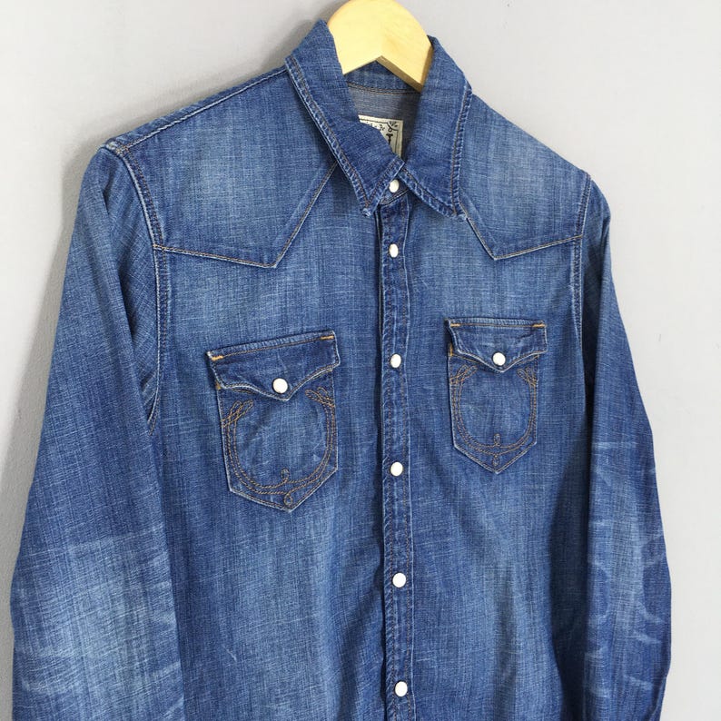 May include: A blue denim shirt with long sleeves, two chest pockets, and a button-down front. The shirt has a classic western-style yoke design and is hanging on a wooden hanger.