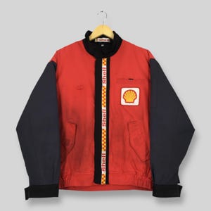 May include: A red and black Shell branded jacket featuring a high collar, long sleeves, and a front zipper closure. The jacket displays the Shell logo on the left chest and a vertical strip with the Shell name and a yellow and red checkered pattern.