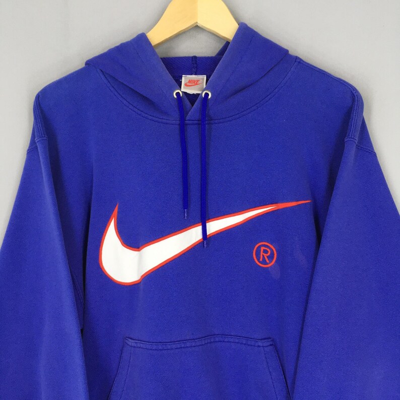 May include: Royal blue hoodie with a white and red Nike logo on the front. The hoodie has a drawstring hood and a kangaroo pocket. The Nike swoosh is outlined in red. The registered trademark symbol is on the right side.